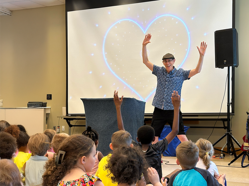 Tom Knight leading an interactive school assembly, with elementary students raising their hands and participating during a music and puppet program