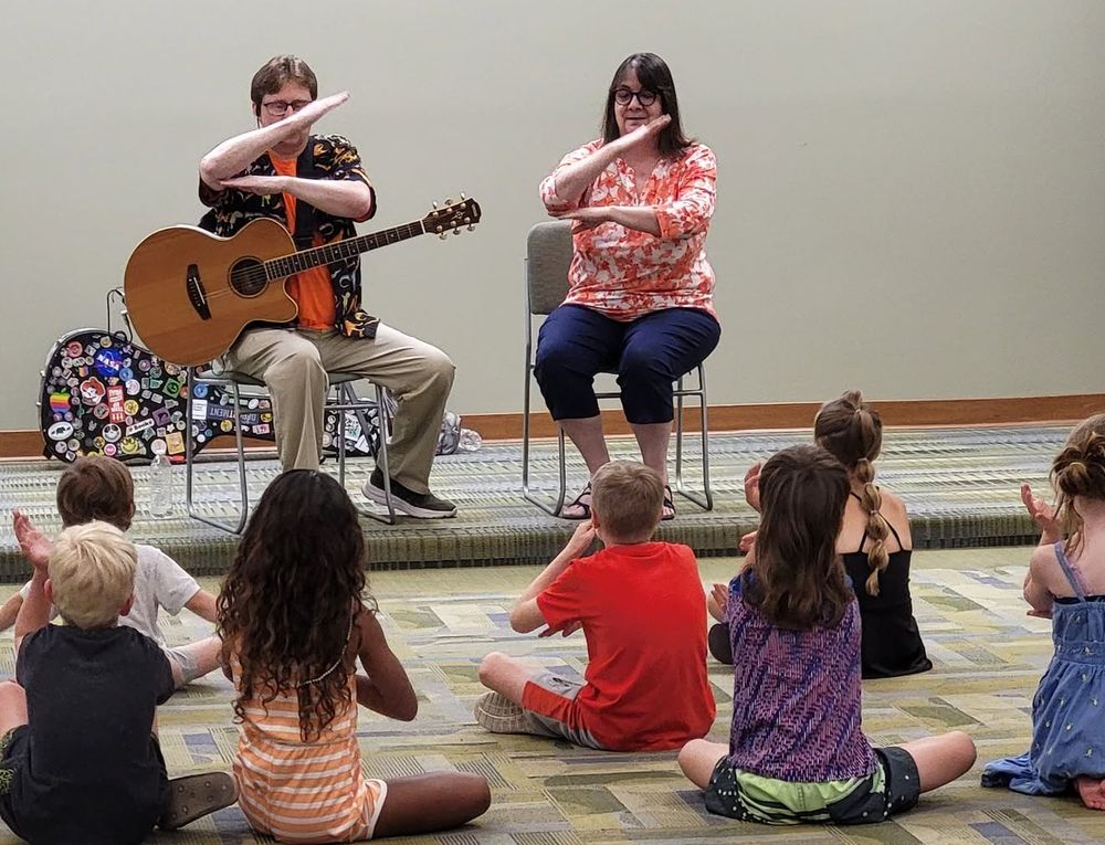 Monty and Lisa demonstrate sign language for "The Cat Came Back" for a crowd of kids at Derby Public Library, July 2026.