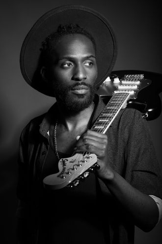 Black and white portrait of Almyr Jules holding an electric guitar, wearing a hat.