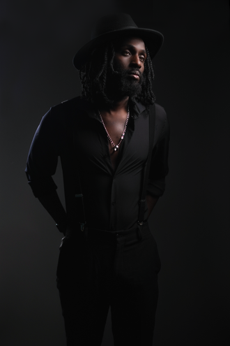 Almyr Jules standing in dramatic single-light portrait wearing black shirt and hat, Soul-Blues Canadian artist promotional photo.