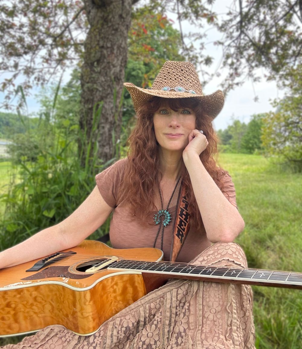 Acoustic guitarist seated outdoors in the Adirondack region of Upstate New York with an acoustic guitar and western hat, representing Traditional Country • Americana • Bluegrass