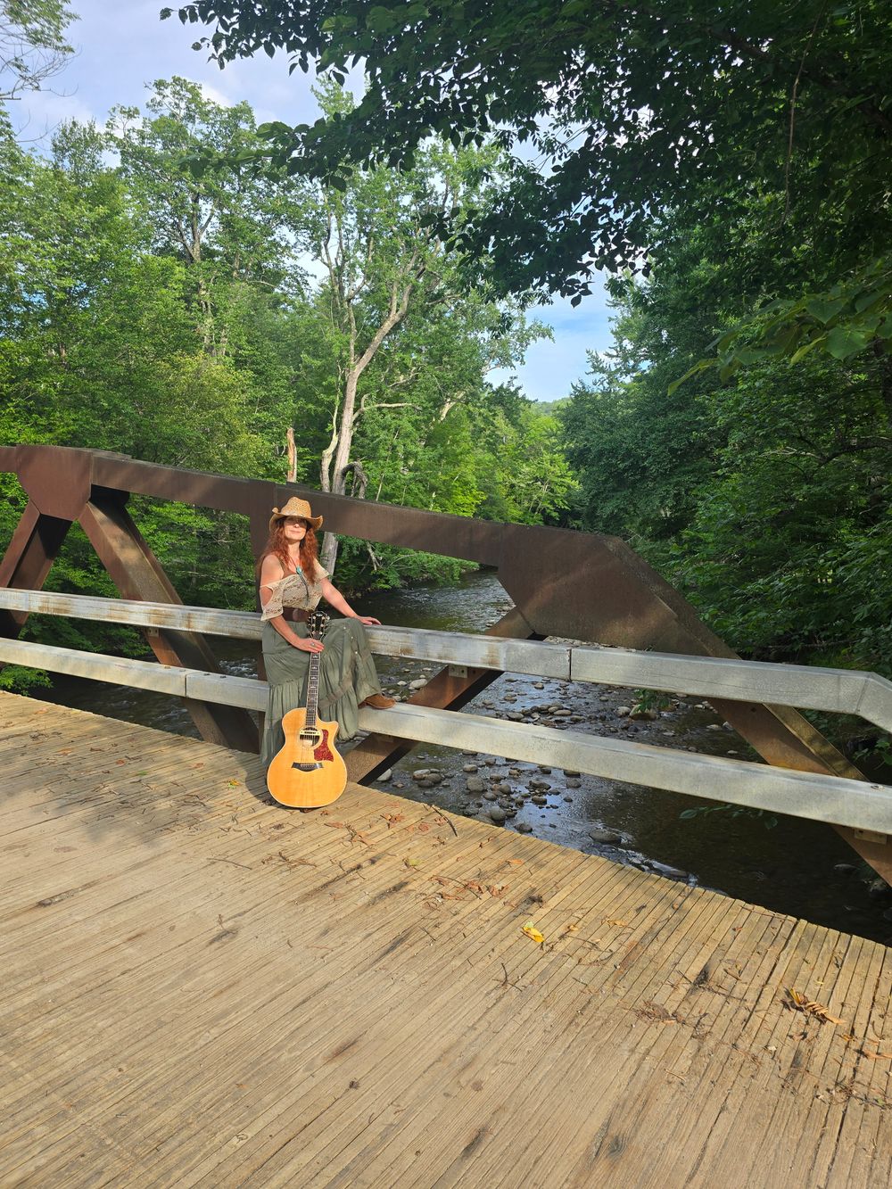 Heather Richards with an acoustic guitar on a rustic bridge overlooking a tree-lined creek in the Adirondacks