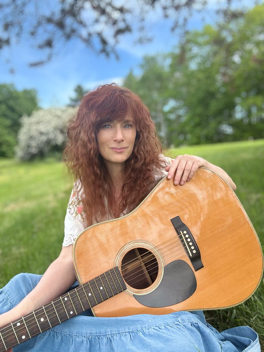 Heather Richards seated outdoors in a grassy field holding an acoustic guitar, wearing a white embroidered blouse and blue skirt with trees and soft greenery in the background.