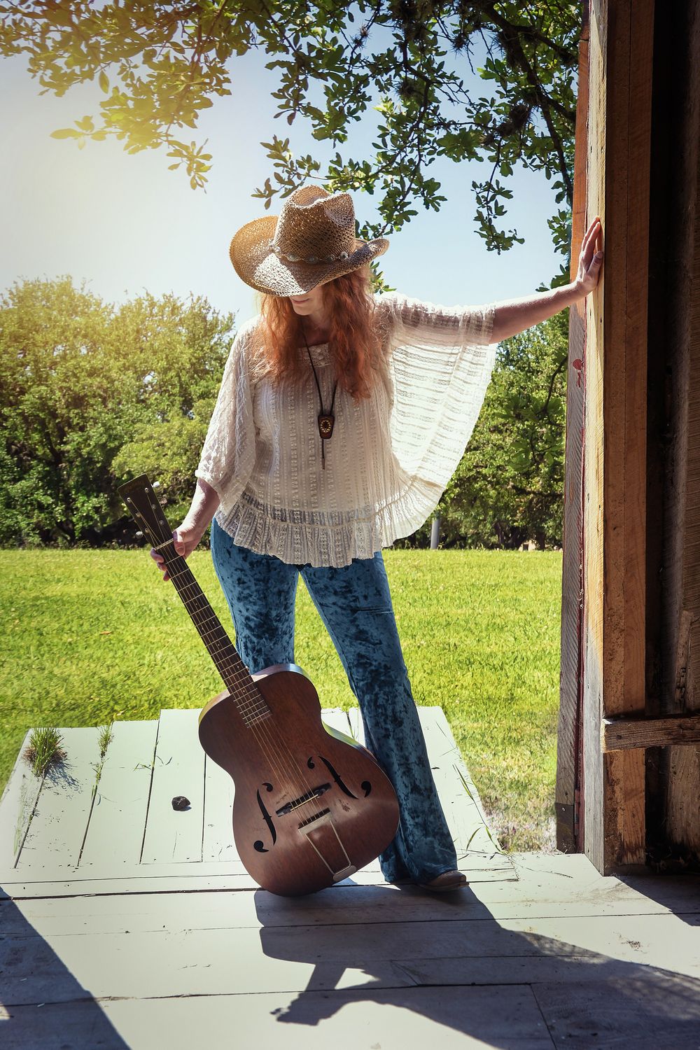 Heather Richards stands on a rustic porch in a cowboy hat, white blouse, and jeans, holding a guitar in the summer sunlight