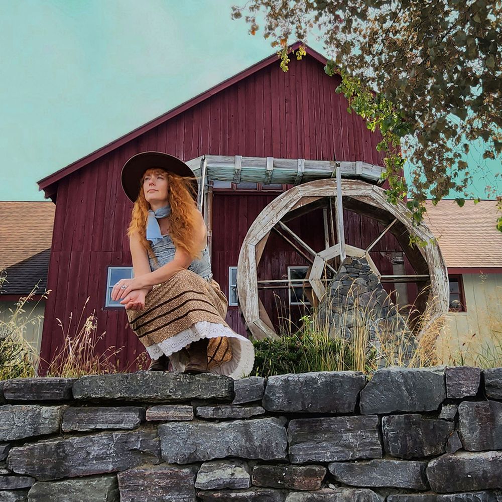 Heather Richards seated on a stone wall beside a rustic water wheel and red barn in an Adirondack-inspired setting, photographed in a warm Americana style.