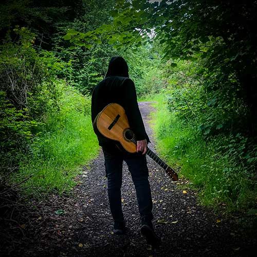 Pete Kavanagh standing with his back to the camera holding an acoustic guitar in a woods