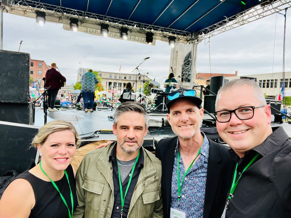 Group photo of Hadrian’s Wall featuring Ashley MacLeod-McRae, Ewen McIntosh, Richard Irwin and Neil Emberg at the Syracuse Irish Festival with the entertainment stage in the background.