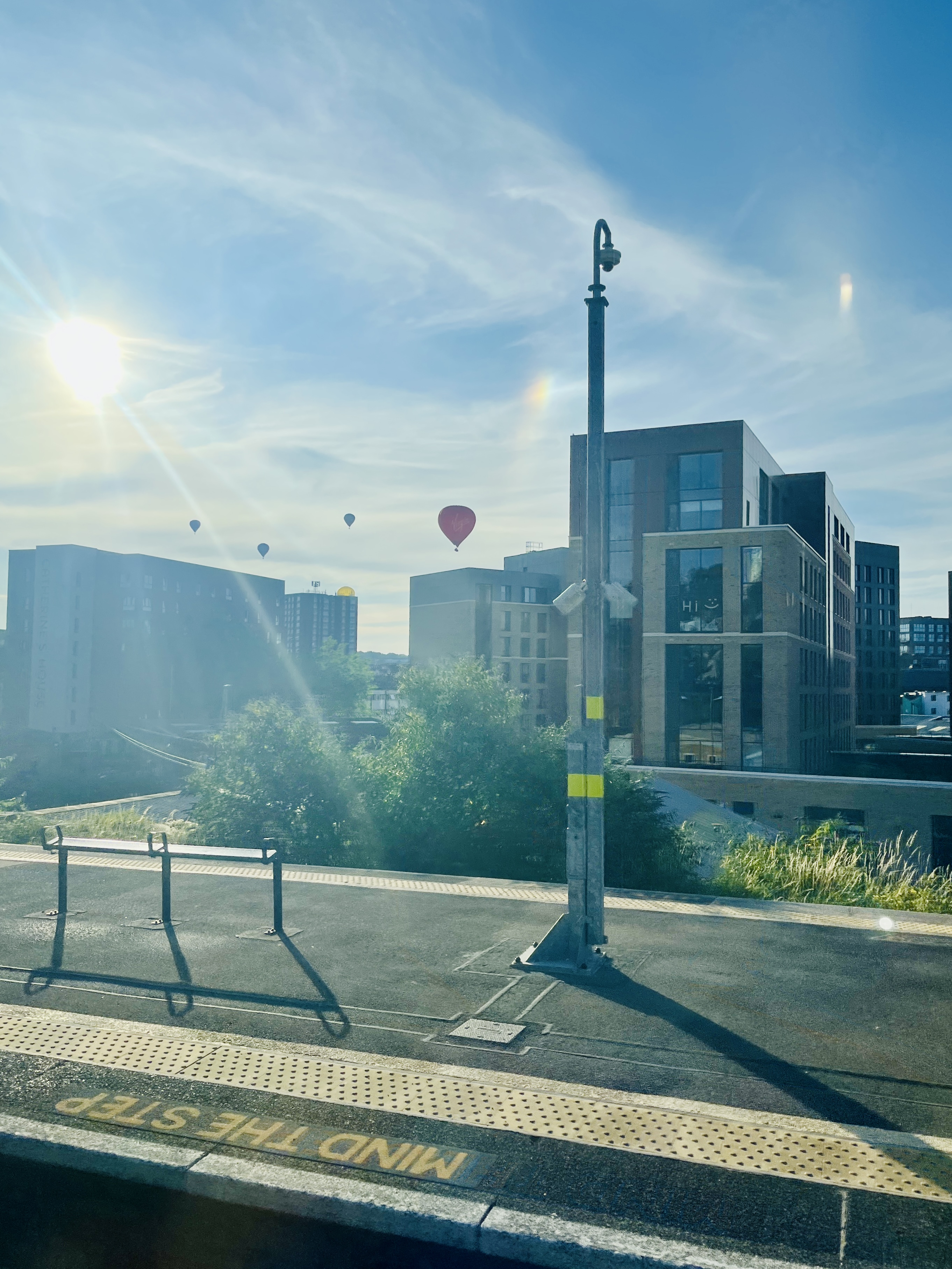 the picture is of some bristol balloons, taken from bedminster train station while on the train, the sky is blue and there is a low strong sun and a mind the step sign on the platform with buildings in the background
