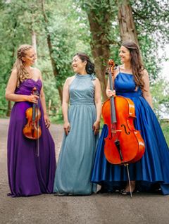 Image: The Champlain Trio, a white woman with long blond hair  holding a violin, a smiling Asian woman in a teal gown, and a white woman with long brown hair holding a cello in a forest