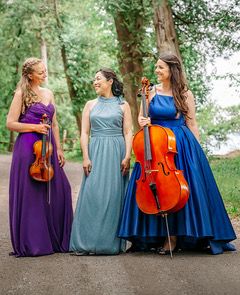 Image: Champlain Trio, a white woman with long blond hair holding a violin, an Asian woman smiling and looking at her and a white woman with long brown hair holding a cello in front of trees