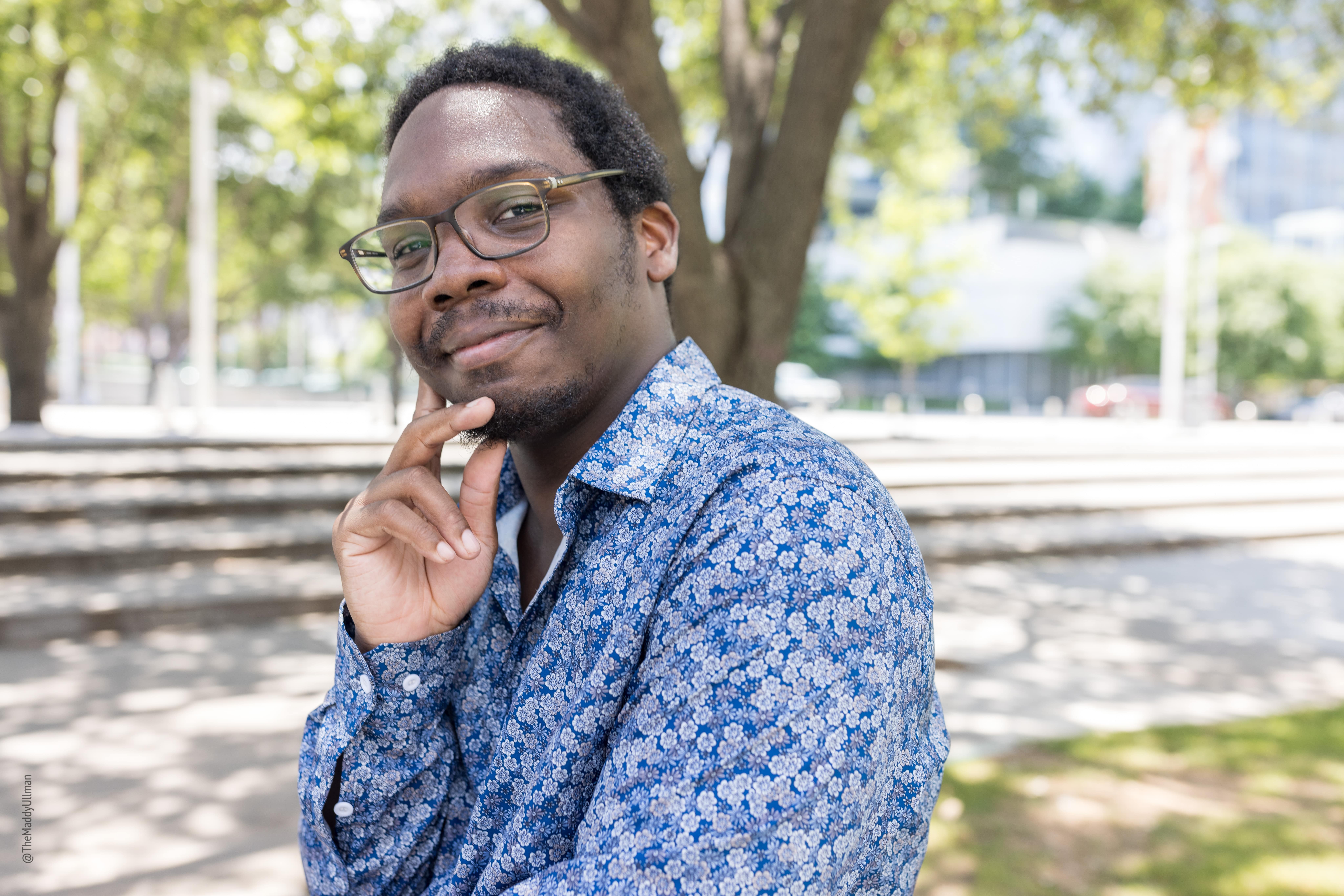 Composer Quinn Mason, a smiling Black man with glasses and a close-trimmed beard and mustache, wearing a blue floral-patterned shirt in a sunny park.