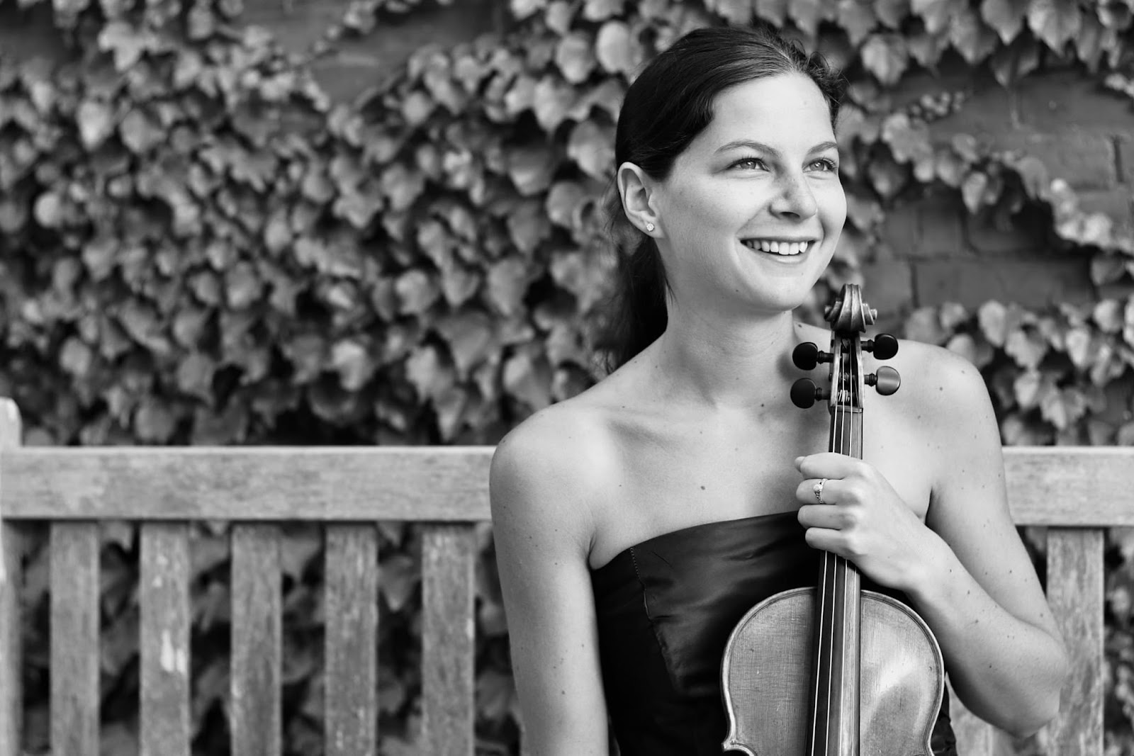 Olivia Moaddel, a smiling white woman in a strapless dark dress holding a violin by the neck while sitting on a bench