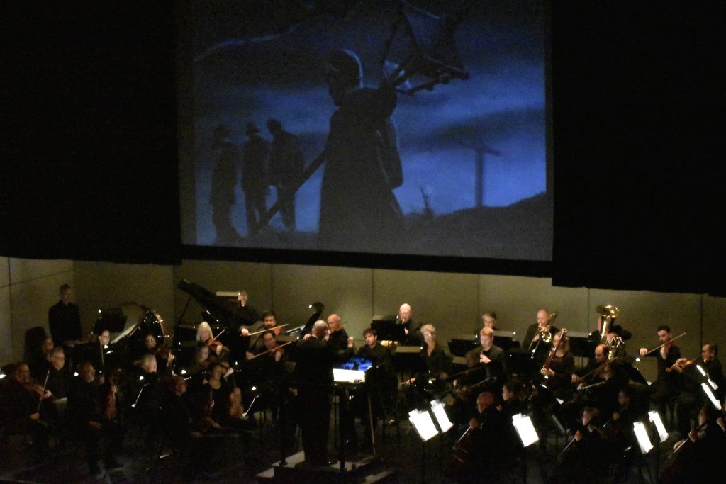 An orchestra in black plays on a stage while a black-and-white film is projected on a screen above them.
