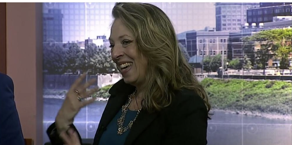 Julia Grella O'Connell, a smiling white woman lifting her hand in front of a background of urban Binghamton