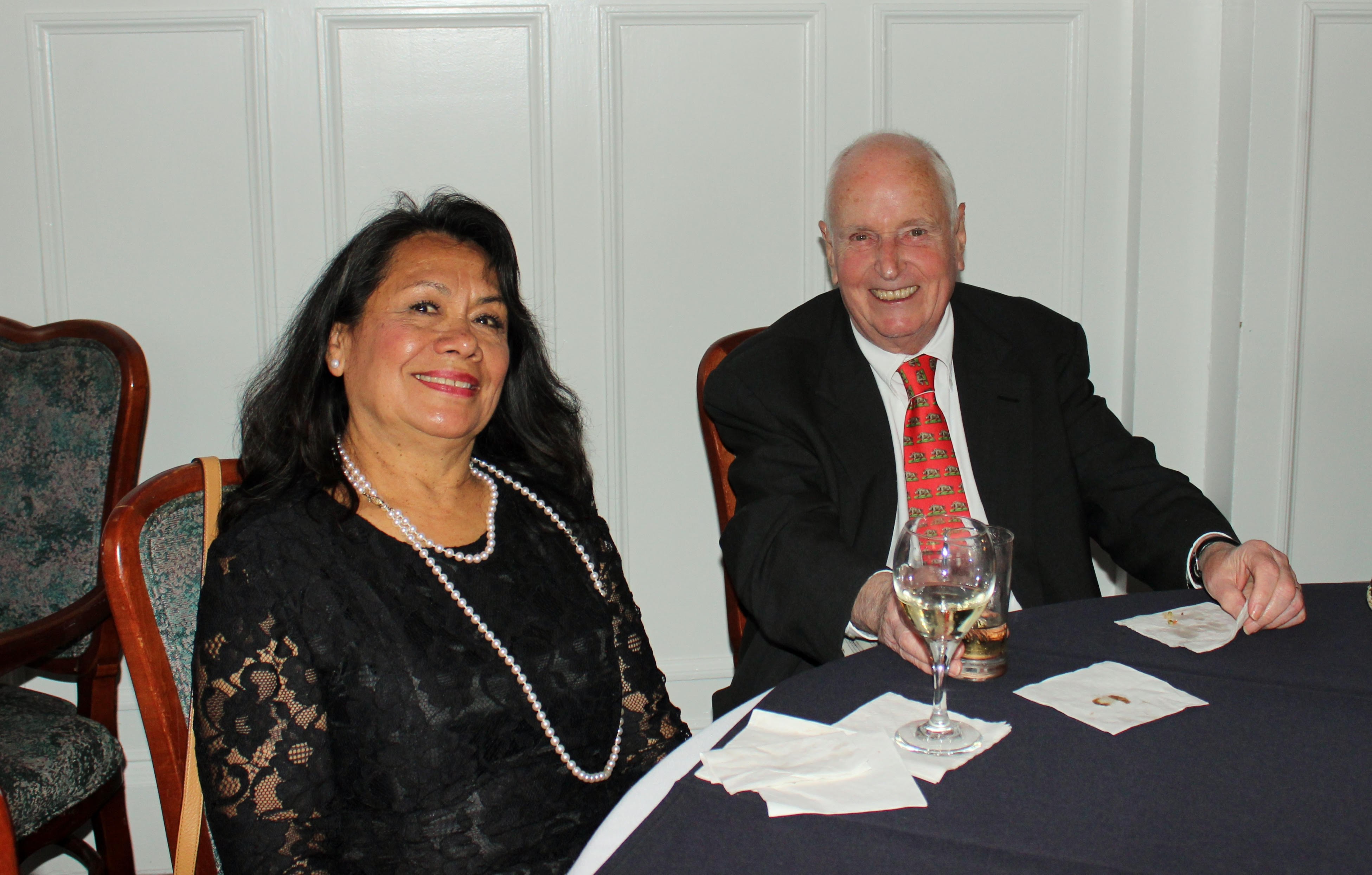 Teresa Babcock, a smiling Latina woman with long black hair and a pearl necklace, and Charlie Babcock, a smiling balding white man with a black jacket and red and green tie, sitting at a table with a glass of wine 