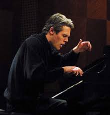 Andrew Russo, a white man with gray hair playing piano with both hands raised over the keyboard and mouth opening concentration