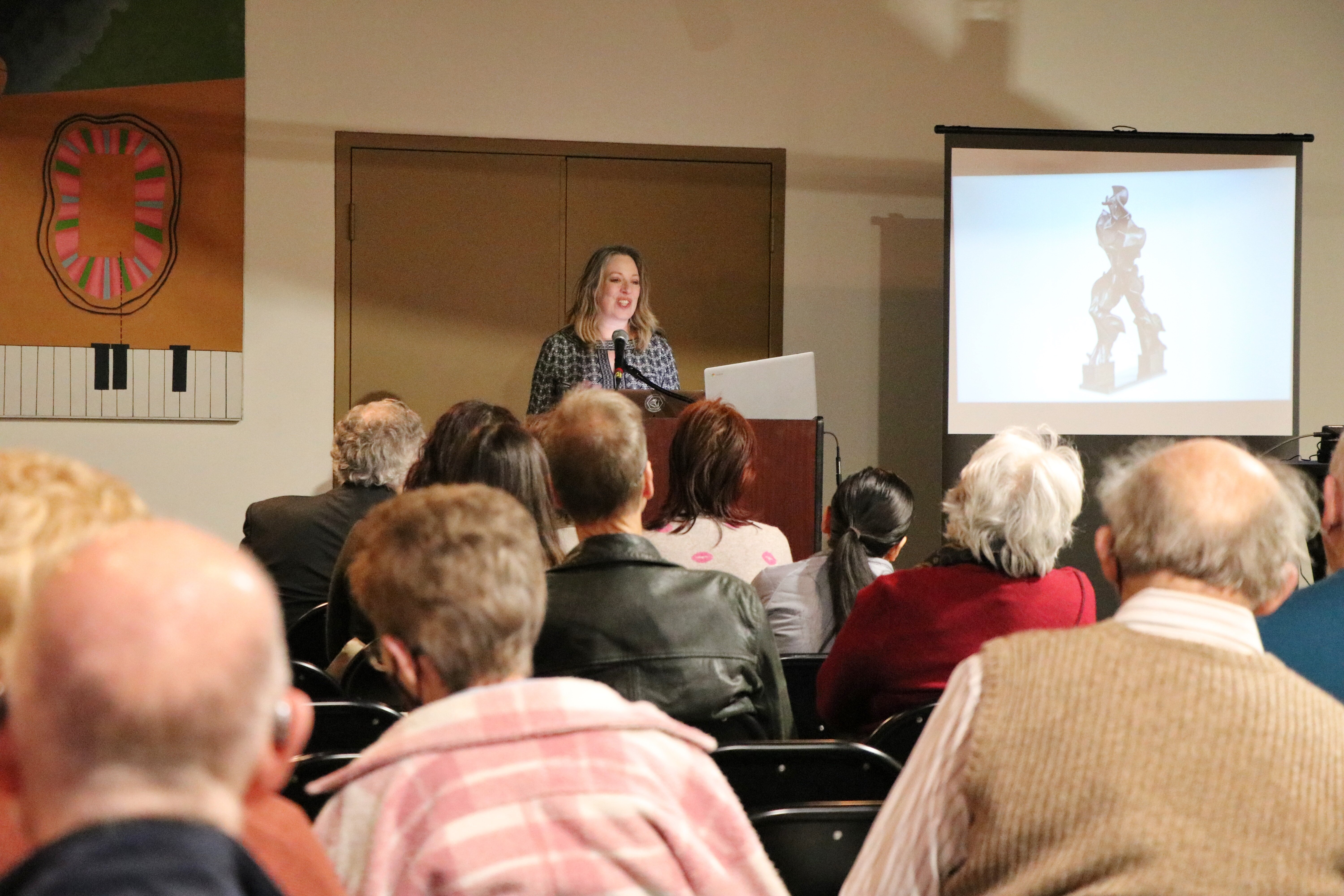 Julia Grella O'Connell, a smiling white woman with shoulder-length brown hair, at a podium in front of an audience at the Broome County Forum Theatre