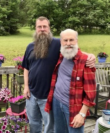 David Williams, a white man with a long brown beard and glasses with his arm around the shoulder of Joseph Varga, a white man with a bushy white beard, standing ion a wooden deck with flowerpots with a yard with trees in the background