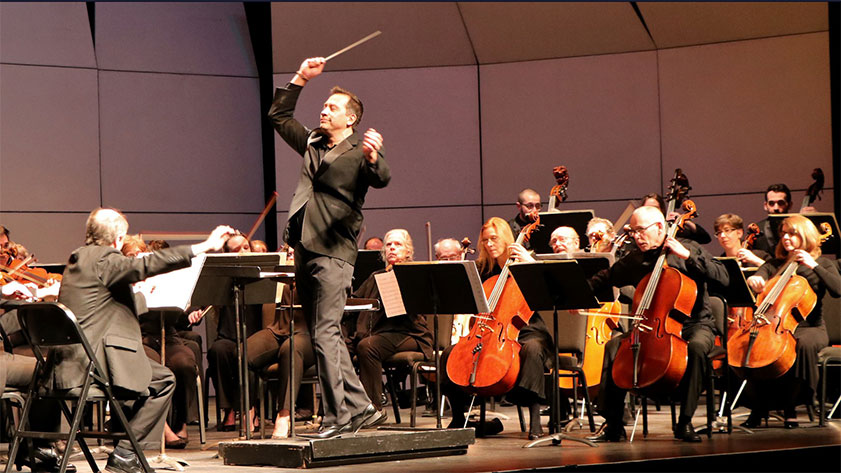 Image: Daniel Hege, a Native American man in a black suit, conducting an orchestra with his baton raised