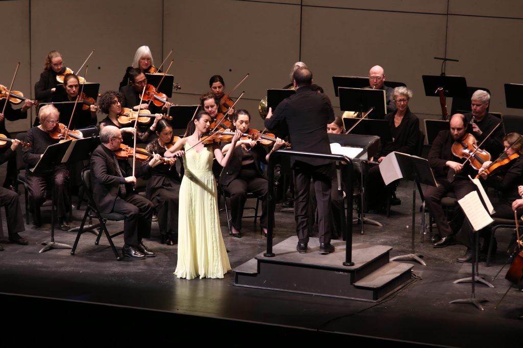 An Asian woman in a long yellow dress playing violin with an orchestra and a conductor in black