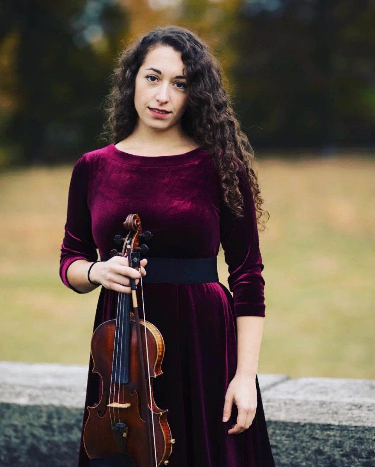 Emily Garrison, a white woman with curly long brown hair in a burgundy velvet gown holding a violin and bow