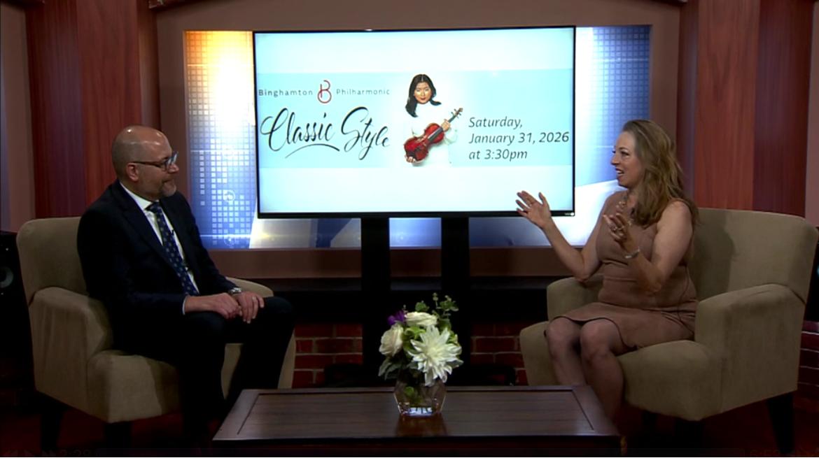 Paul Cienniwa, a bald white man with glasses, and Julia Grella O'Connell, a white woman with long brown hair and hands raised, sitting in beige chairs talking in front of a screen showing an image of violinist Hina Khuong-Huu, an Asian woman in white holding a violin