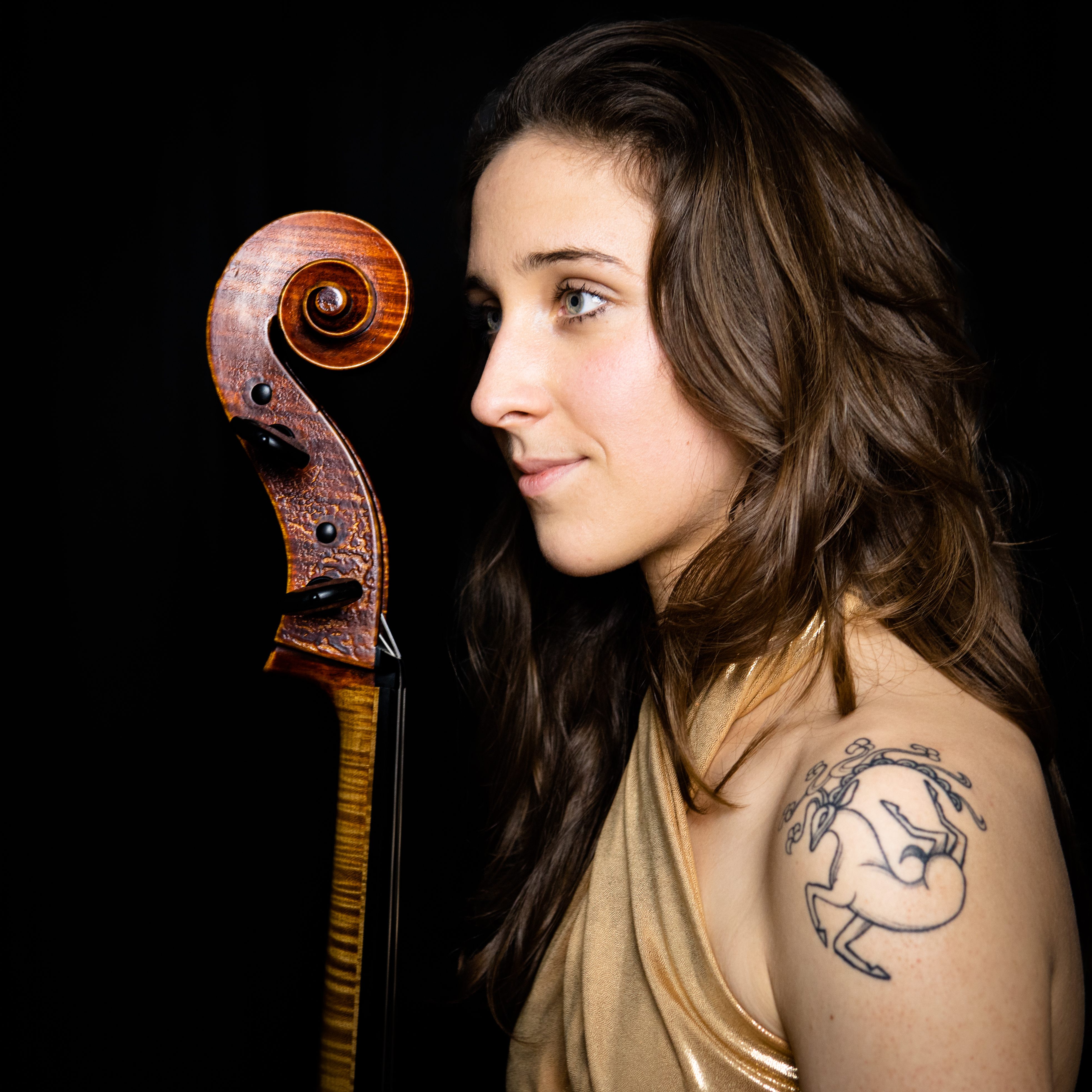 Annie Jacobs-Perkins, a white woman with long brown hair shown in profile, with a tattoo on her exposed shoulder of a Siberian reindeer, holding the neck of a cello