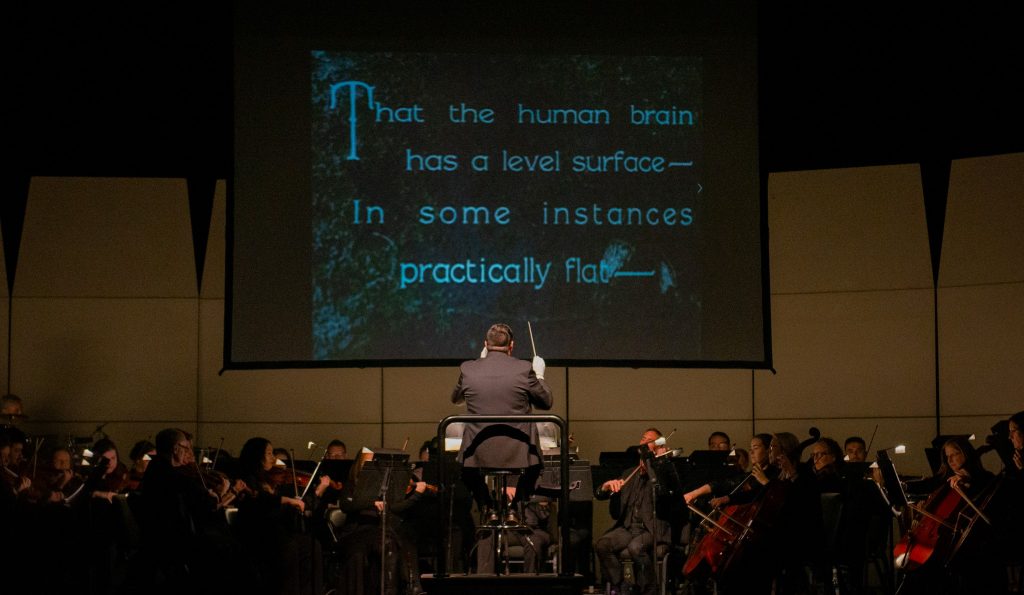 dark image of the orchestra with conductor; a movie screen is above them with an image from a silent film