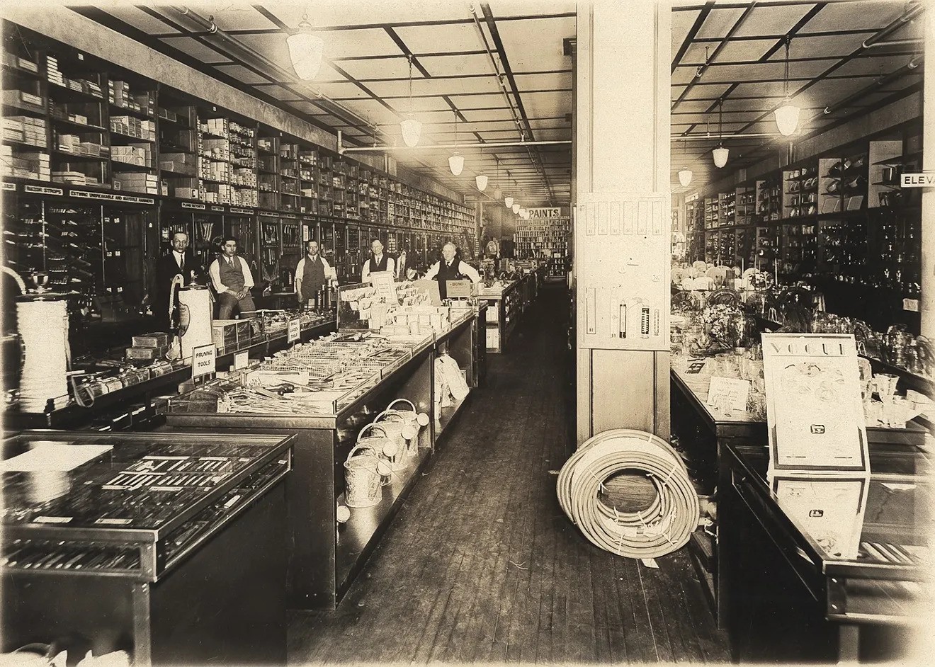 Sepia-toned black and white photo of old-fashioned hardware store with wooden floors, hardware items laid out on broad counters with six male employees in shirt sleeves, waistcoats, and ties standing in the rear