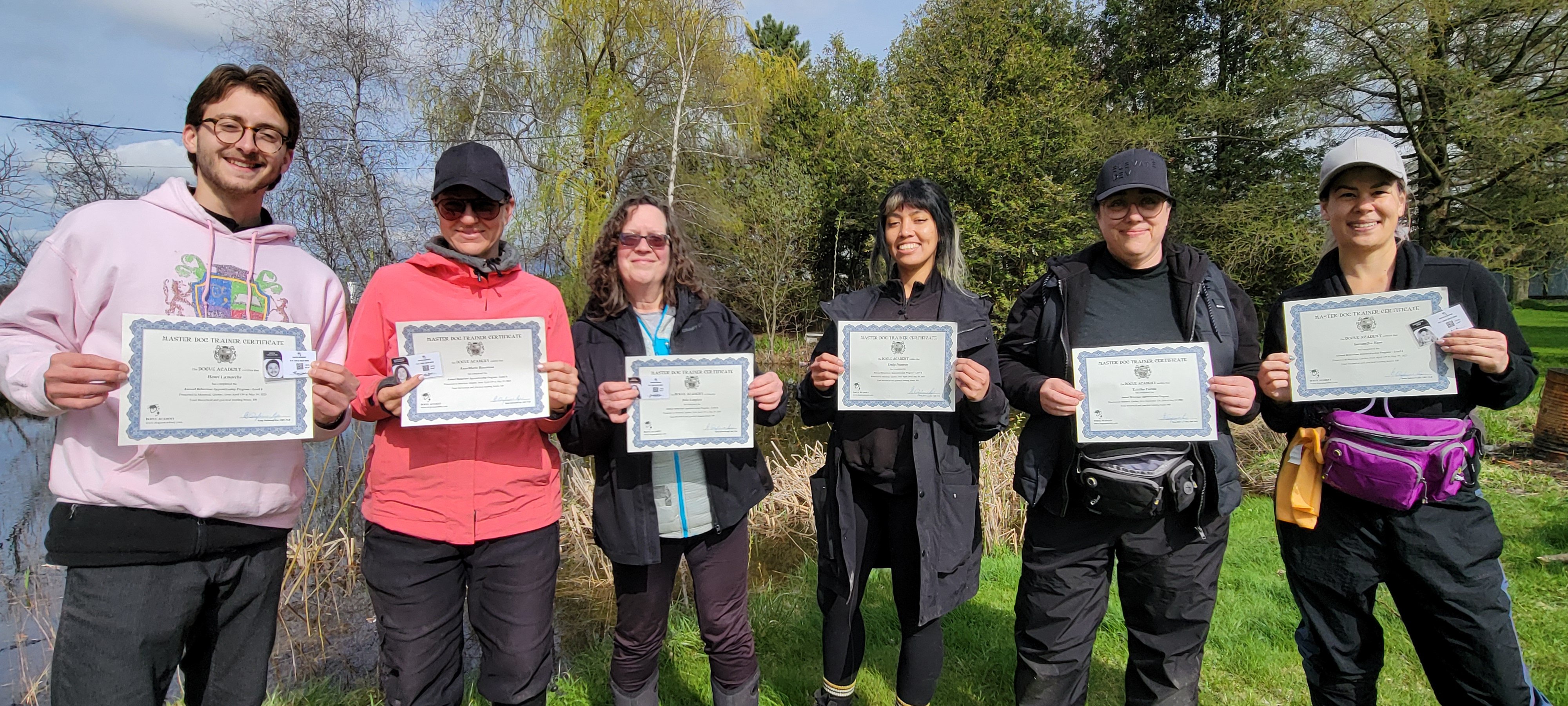 A cohort of students from the 2025 professional dog trainer certification course posing with their certificates in front of a lake