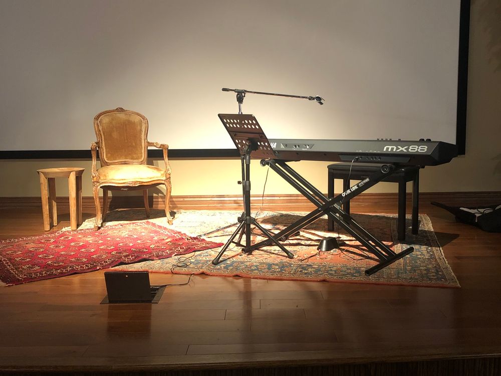 Image of small stage with keyboard, antique french chair and small table on Persian rugs. There is a microphone. This is to illustrate the musical's humble start in Little Rock, Arkansas 2023