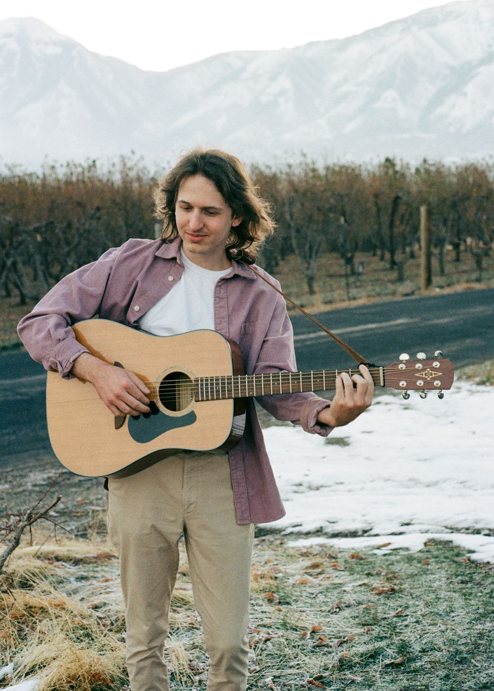Image of Jackson Harden playing acoustic guitar next to a snowy road with snow capped mountains and an orchard of dead trees in the background. Photo by Z Gray Adams.