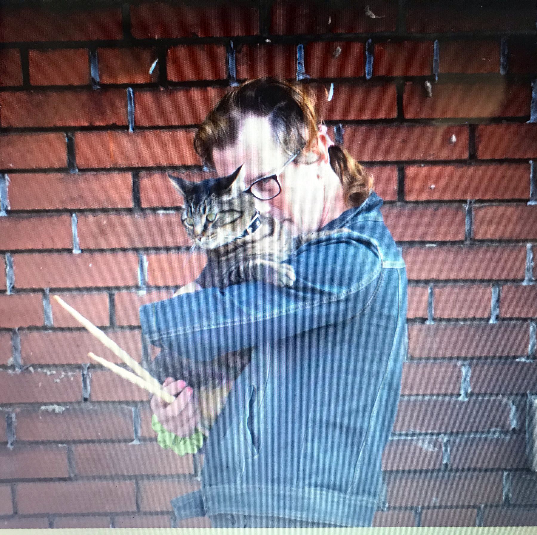 Polly-Jean hugging Dwayne the cat against a brick wall, drumsticks in hand