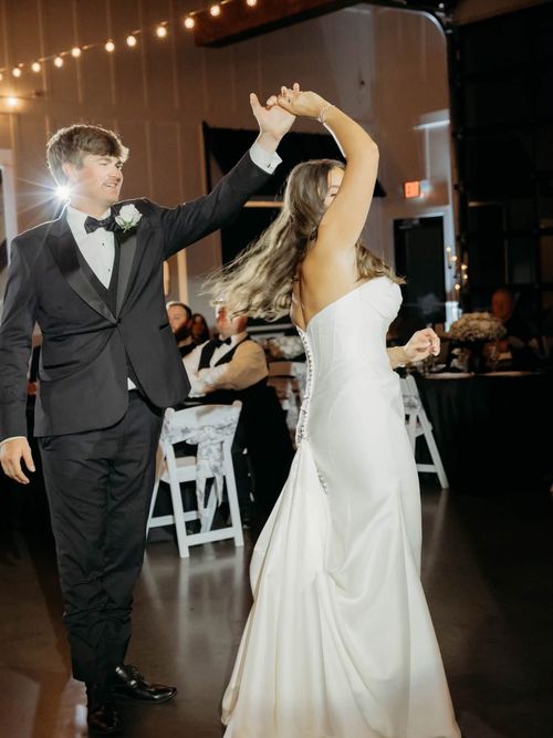 Groom twirling the bride on the dance floor during their wedding reception, with guests and string lights in the background.