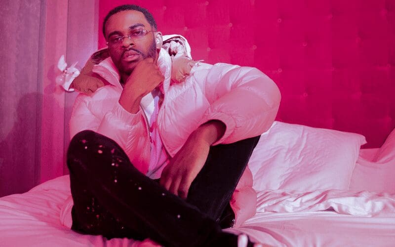 Mista Strange wearing a pink puffy windbreaker, sitting on the bed of a pink bedroom