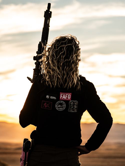 One of the female instructors holding a rifle standing on a hill back towards the camera in front of a sunset