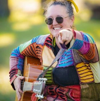Jen Anders smiling with her acoustic guitar outdoors.
