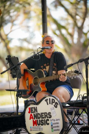 Jen Anders playing acoustic guitar at an outdoor live show.
