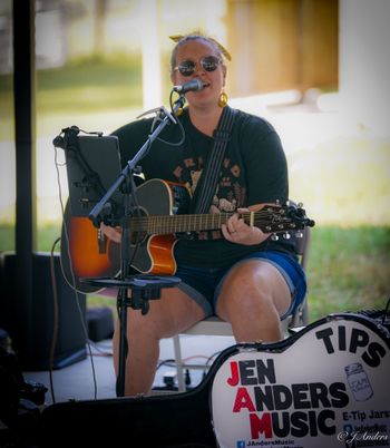 Jen Anders performing seated with an acoustic guitar and microphone.
