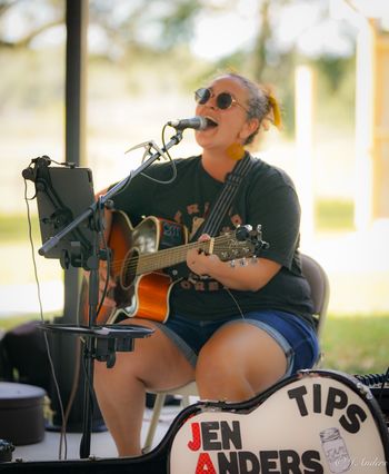 Jen Anders singing and playing acoustic guitar outdoors.
