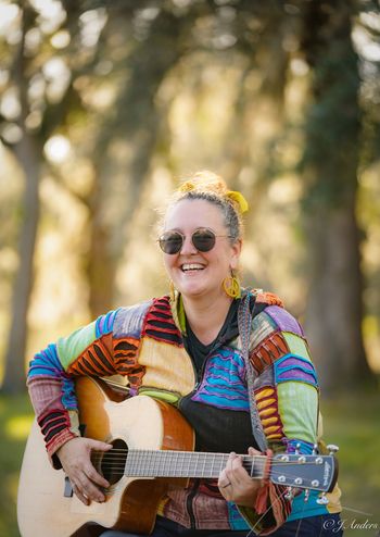 Jen Anders smiling with her acoustic guitar in an outdoor portrait.
