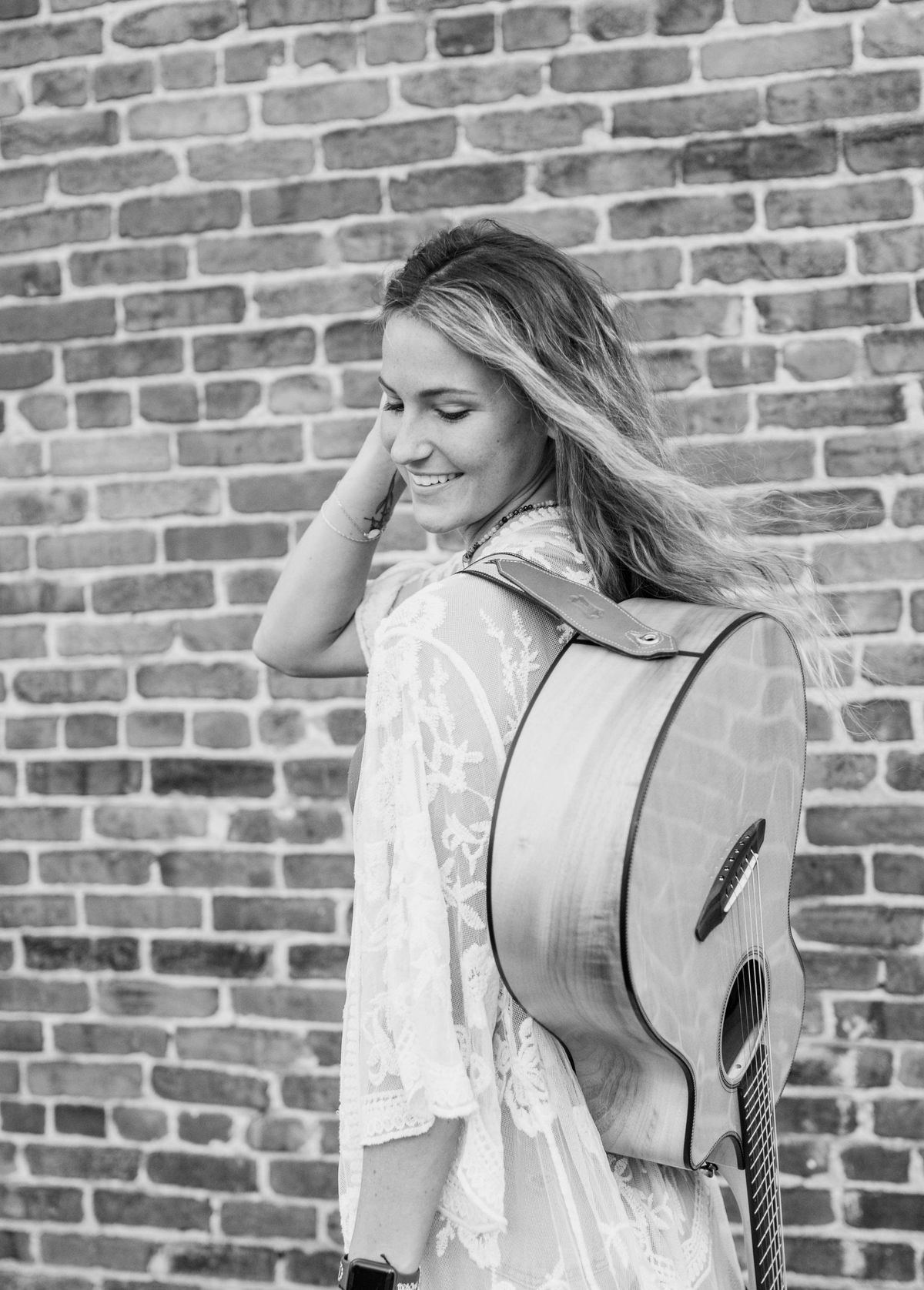 Holland Dotson in black and white, guitar case on her back against a brick wall