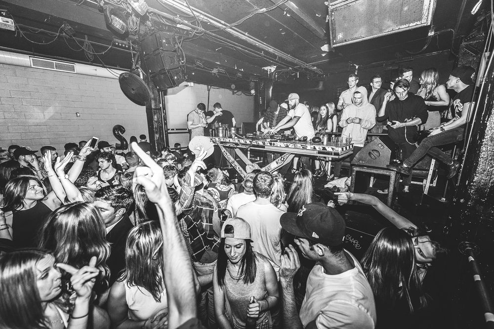 A group of young people dancing and drinking in a warehouse listening to a DJ paying music. The photo is in black and white.