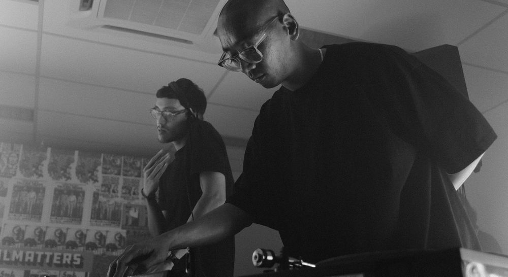 Two DJs setting up a record on a turntable. One of them is listening to another record on headphones.